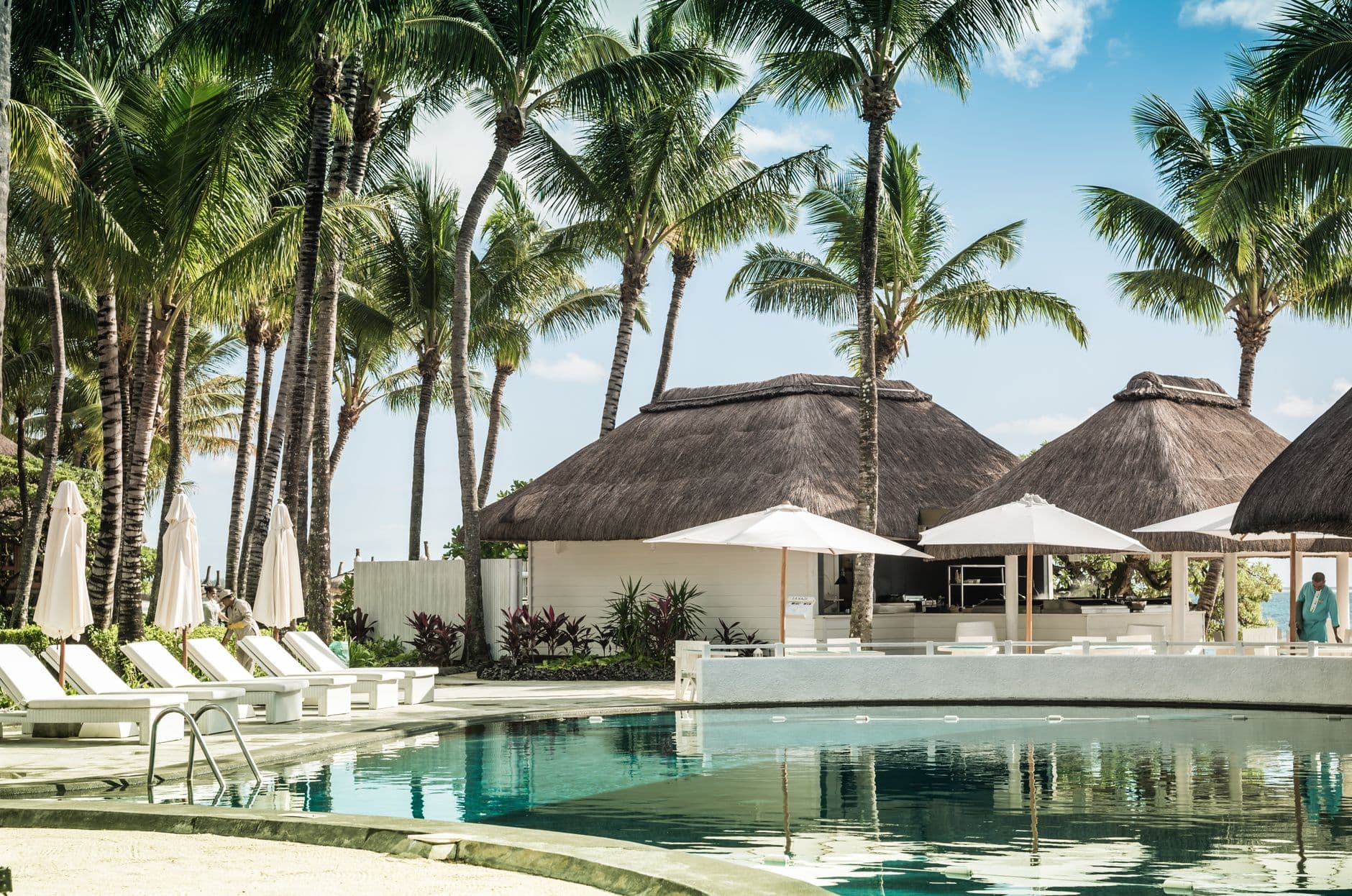 Een zwembad met ligbedden, palmbomen en rieten daken bij een resort