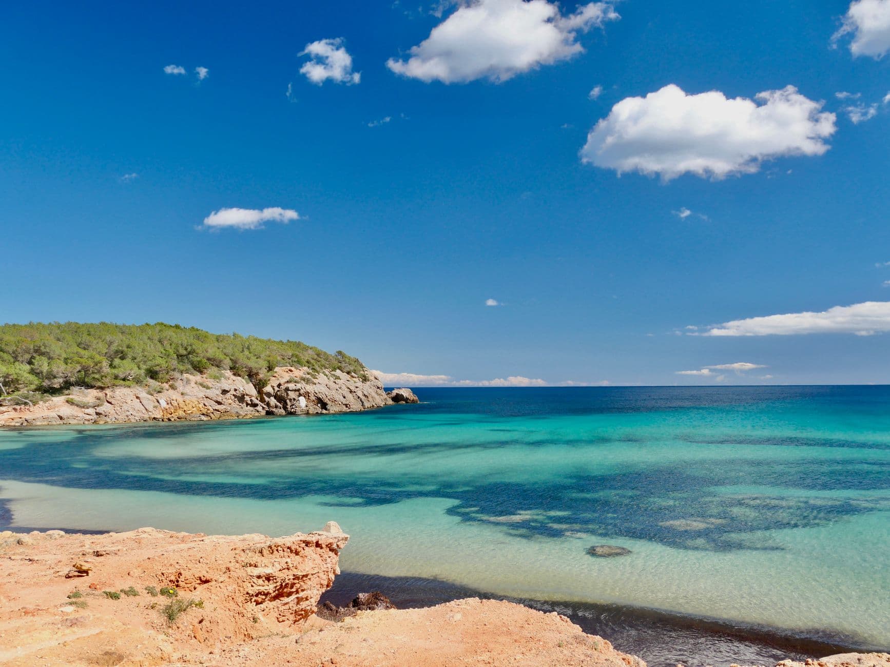 Een baai met turquoise water tussen rode rotsen en een groene rotskust, onder een blauwe lucht op Ibiza