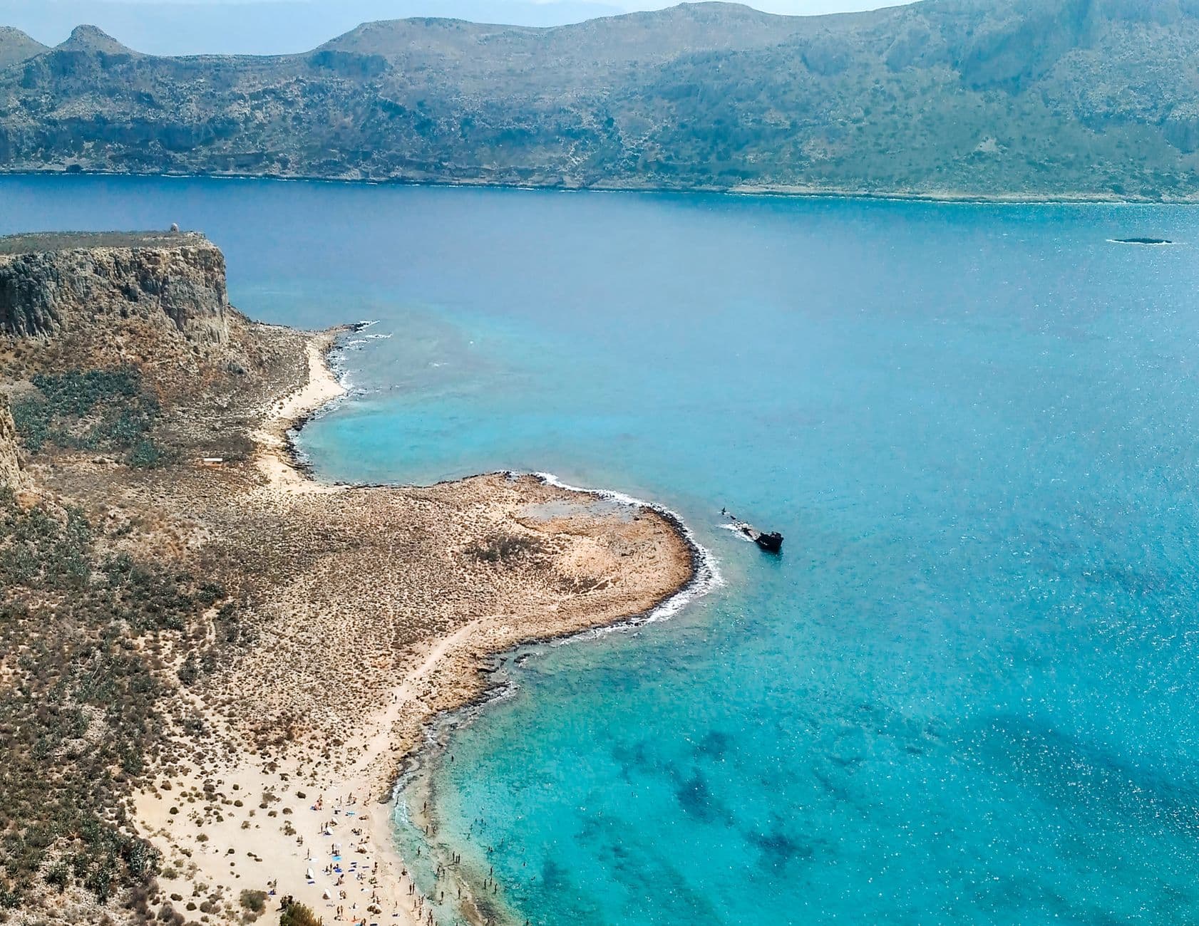 Luchtbeeld van een strand op Kreta met turquoise water, een hoge rotskaap en een scheepswrak voor de kust