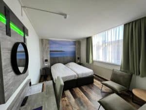 Interieur van een hotelkamer met twee opgemaakte eenpersoonsbedden, twee groene fauteuils, een bureau, een grote houten muurdecoratie met een rond gat erin, groene gordijnen en een raam.