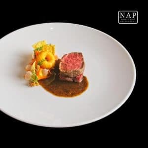 Wit, rond bord met een stuk gebakken vlees, een gefrituurde ring, groenten, saus, en het logo van "NAP HOTEL CAFÉ RESTAURANT" in de rechterbovenhoek. De achtergrond is zwart.