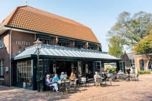 Foto van Hotel Restaurant De Jong, een gebouw met donkerkleurige gevel, grote ramen, een gele luifel en een terras met tafels en stoelen. Straatnaambord "Katteijnestraat" is zichtbaar in de rechterbovenhoek.