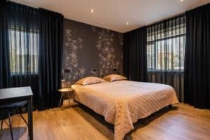 Een hotelkamer met een tweepersoonsbed met een licht dekbed, twee ramen met zwarte gordijnen, een behang met een bloemenpatroon op de muur achter het bed, een kleine ronde tafel met een lamp, en een bureau met stoel op de voorgrond.