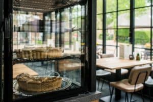 Een glazen vitrine met twee appeltaarten op zilveren schalen. Op de achtergrond is een restaurantinterieur te zien met een houten tafel en stoelen, grote ramen en groen buiten.